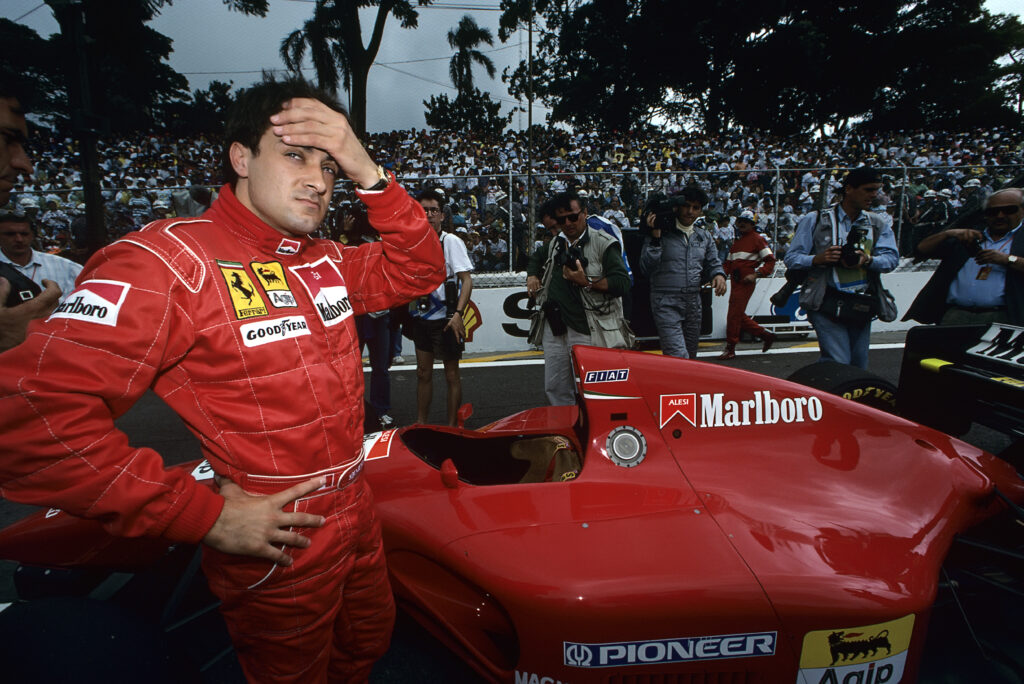 Jean Alesi stands next to his Ferrari on the grid with a hand on his head.